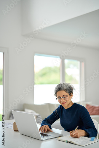 Mature 50 years old lady wearing glasses remote working online on computer in living room. Happy middle aged woman sitting at table at home office using laptop learning online writing notes. Vertical