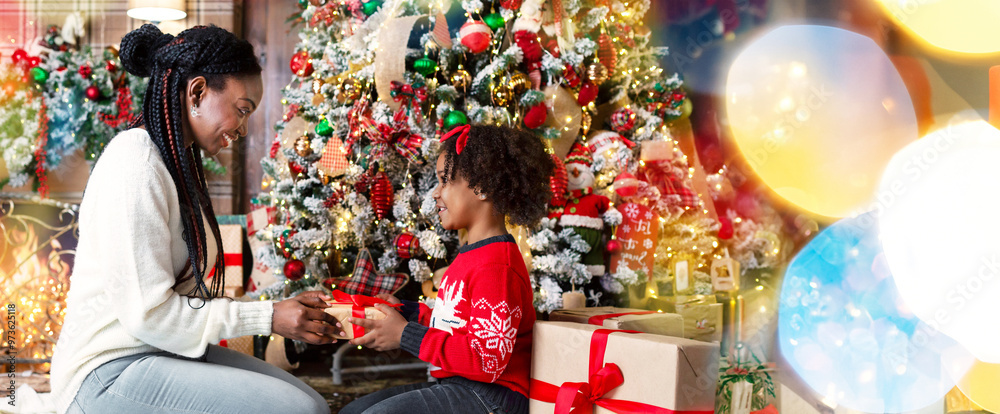 Merry Christmas. African american mother and daughter exchanging gifts sitting under Xmas tree at home, side view Merry Christmas. African american mother and daughter exchanging gifts sitting under Xmas tree at home, side view