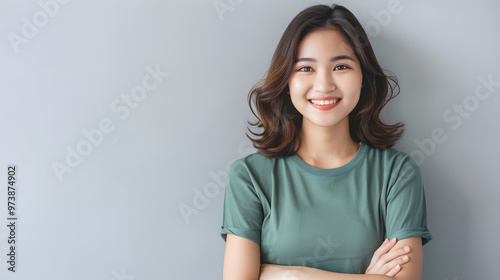 Confident young Asian woman with shoulder-length wavy hair and a bright smile, wearing a green t-shirt, arms crossed, against a light gray background