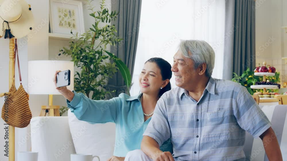Old Asian couple spending leisure time at home using a phone for online entertainment. Positive and happy moment shows enjoyment of the internet and bonding while resting on the soft sofa Old Asian couple spending leisure time at home using a phone for online entertainment. Positive and happy moment shows enjoyment of the internet and bonding while resting on the soft sofa
