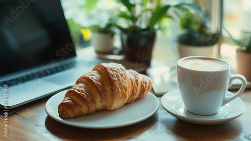 A cup of coffee with latte art and a croissant on a white plate, placed on a wooden desk with a laptop and potted plants nearby.