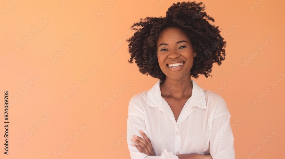 A happy, young black woman with curly black hair smiles while standing in front of a peach - colored backdrop, wearing a white button - up shirt with her arms crossed and is lightly playing with her A happy, young black woman with curly black hair smiles while standing in front of a peach - colored backdrop, wearing a white button - up shirt with her arms crossed and is lightly playing with her