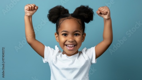 A little Black girl with a big smile and her fists raised in the air celebrates a victory or achievement. She is wearing a white shirt and her dark hair is styled in a high ponytail, copy space photo