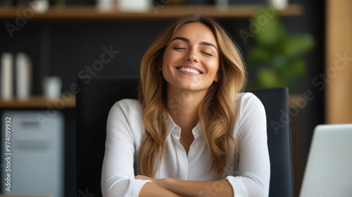 A woman in a white shirt smiles peacefully with eyes closed, sitting at her office desk, enjoying a quiet, relaxed moment.