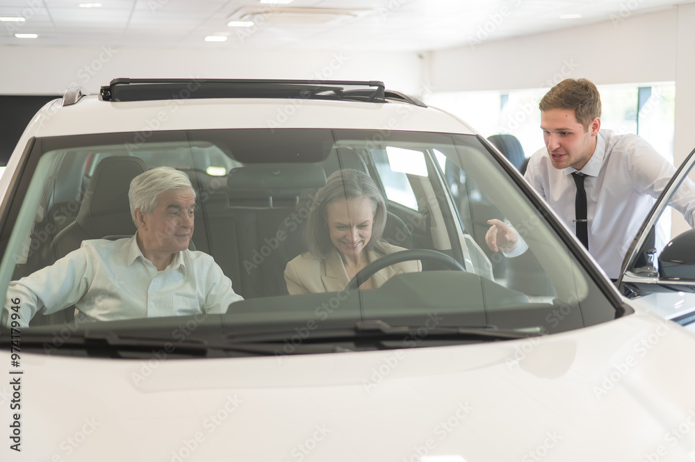 A gray-haired Caucasian woman is sitting behind the wheel of a car. Car dealership salesman consulting mature married couple. A gray-haired Caucasian woman is sitting behind the wheel of a car. Car dealership salesman consulting mature married couple.