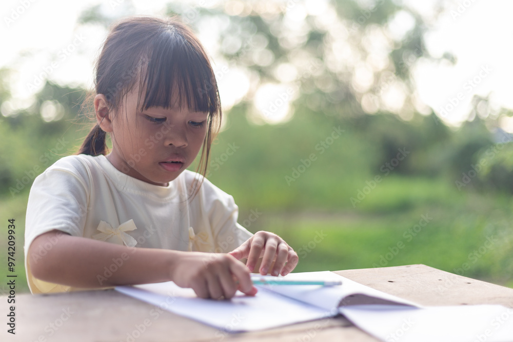 A young girl is sitting at a table writing. She is wearing a white shirt and has her hair in a ponytail. A young girl is sitting at a table writing. She is wearing a white shirt and has her hair in a ponytail.