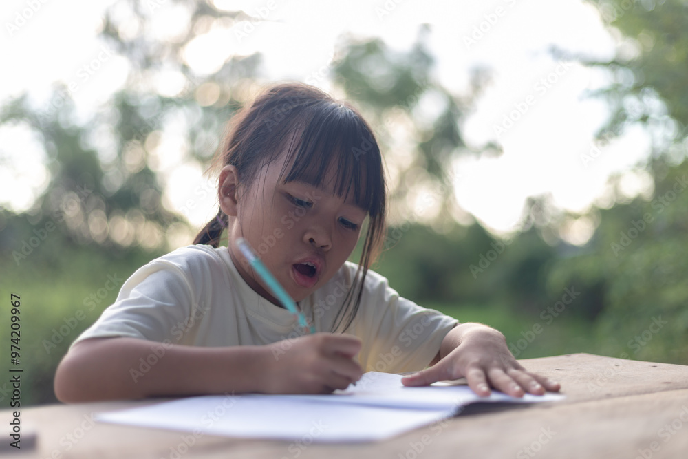 A young girl is sitting at a table writing. She is wearing a white shirt and has her hair in a ponytail. A young girl is sitting at a table writing. She is wearing a white shirt and has her hair in a ponytail.