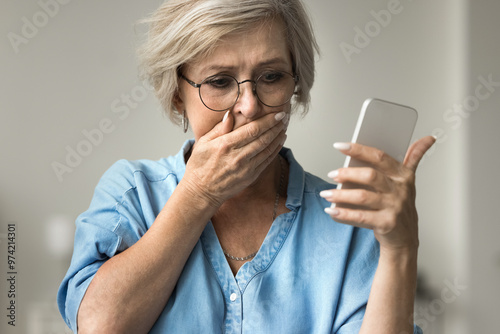 Close up older woman looks at smartphone screen with distressed expression, cover her mouth with hand, read alarming news on phone, feel unhappy, scared. Financial setback, phishing, awful information photo