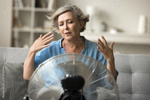 Drained mature woman sits on sofa cooling herself using electric floor ventilator, fan blowing to face of senior female suffers from overheating with eyes closed indoors without air conditioner system