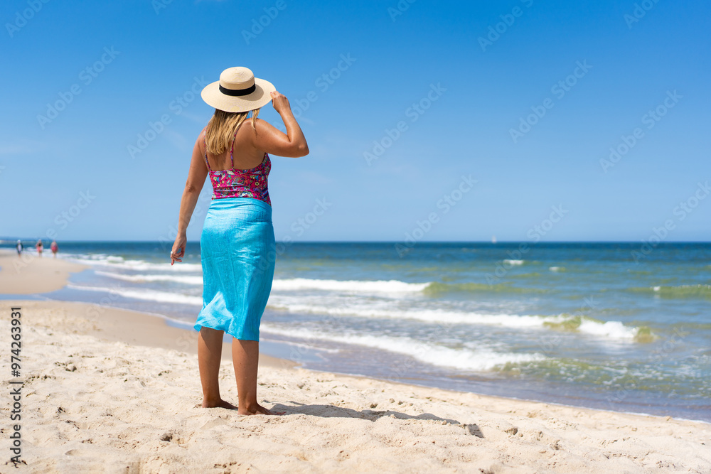 Summer vacation. Tanned woman in colorful swimsuit, blue pareo and sun hat standing by shore on sandy beach on beautiful sunny day. Back view Summer vacation. Tanned woman in colorful swimsuit, blue pareo and sun hat standing by shore on sandy beach on beautiful sunny day. Back view