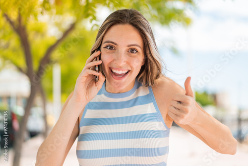 Young pretty woman at outdoors using mobile phone while doing thumbs up