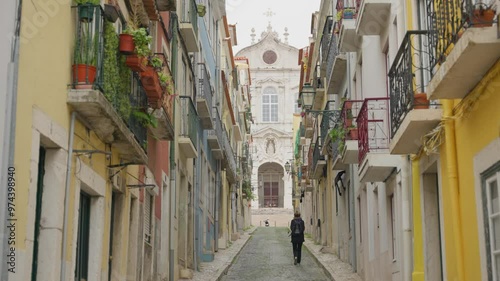 Female tourist or student walking up a street with colorful building in Portugal. Beautiful magical looking architecture in Lisbon. Balconies with flowers and laundry hanging. Church in the background