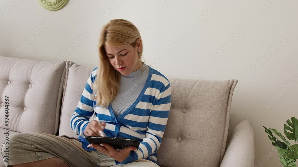 Cute young blond woman in a striped blouse drawing a heart in a tablet with a stylus pen while sitting cross legged on a light couch in a room.