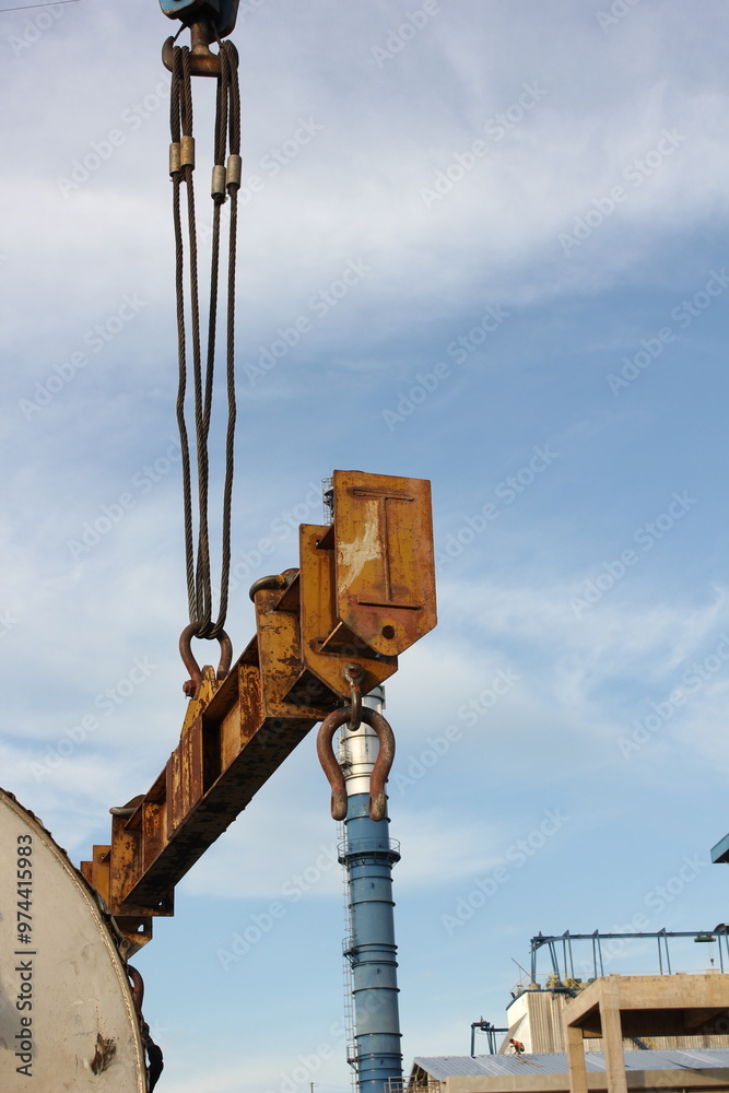 A large, rusty metal beam is being lifted by a crane against a blue sky with white clouds. The beam has hooks attached to it and is suspended by thick cables. An industrial factory with chimneys A large, rusty metal beam is being lifted by a crane against a blue sky with white clouds. The beam has hooks attached to it and is suspended by thick cables. An industrial factory with chimneys