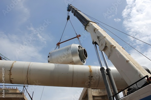 A large crane lifts a cylindrical object onto a long, white cylinder. The crane is white with a blue boom, and the sky is blue with white clouds.