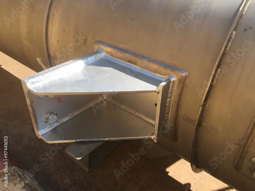 A close-up of a triangular stainless steel support bracket welded to a cylindrical pipe, secured with bolts. Visible weld marks outline the edges where the bracket is attached.