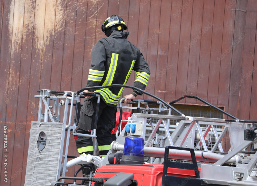 Fototapeta premium Fearless firefighter in uniform walks confidently on the fire truck ladder with a blue siren during an emergency