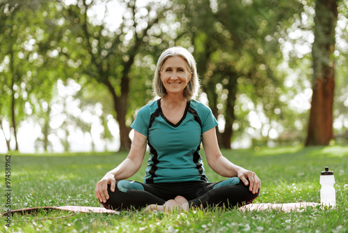 Smiling beautiful senior woman practicing yoga in park sitting on yoga mat, outdoors