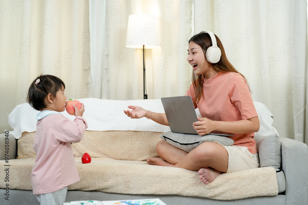 Asian woman working from home, wearing headphones and holding laptop. Engaging with toddler girl who is holding fruit toy. Cozy home environment, balancing remote work and parenting responsibilities.