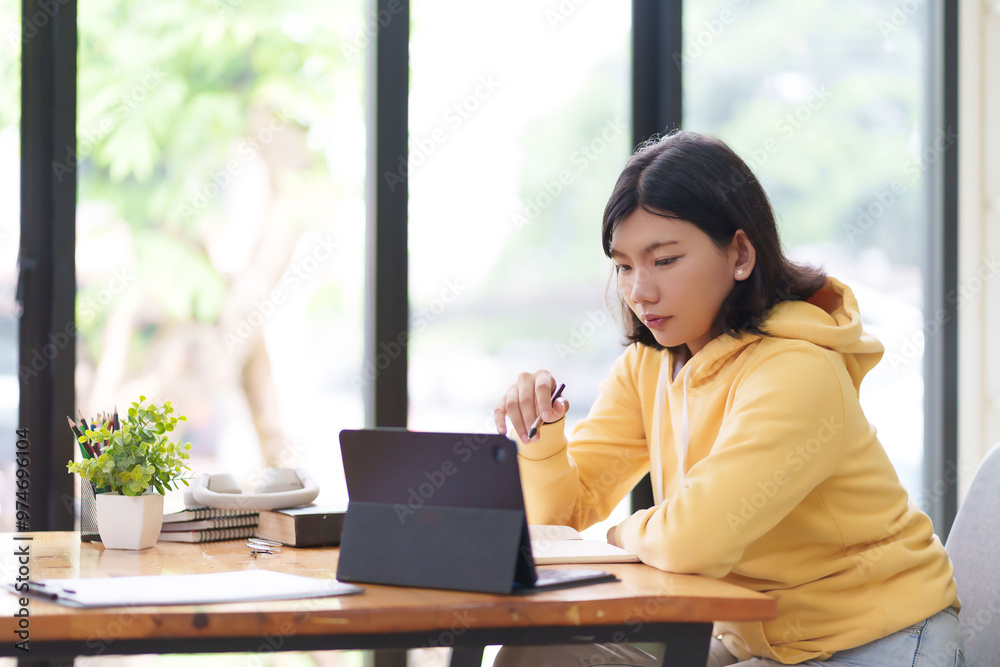 A young woman in a yellow hoodie is sitting at a table, typing on her laptop. She is working in a bright room with large windows, surrounded by natural light and greenery outside A young woman in a yellow hoodie is sitting at a table, typing on her laptop. She is working in a bright room with large windows, surrounded by natural light and greenery outside