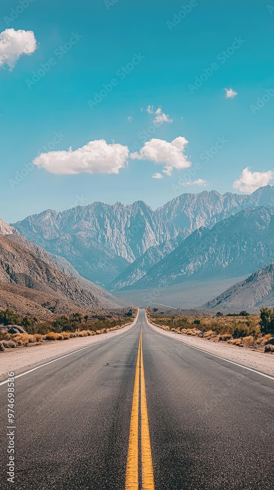 Fototapeta premium A solitary highway stretches towards the horizon, framed by towering mountains and a clear blue sky. The yellow lines guide the way, promising adventure and unknown destinations.