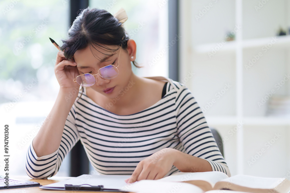 A young woman in a striped shirt is sitting at a desk, resting her head on her hand while working on a notebook. She appears thoughtful, with a plant and an organized workspace around her A young woman in a striped shirt is sitting at a desk, resting her head on her hand while working on a notebook. She appears thoughtful, with a plant and an organized workspace around her