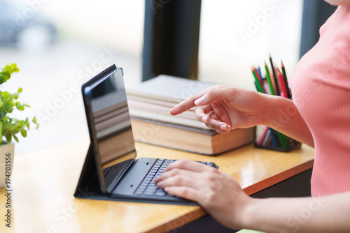 A young woman in a pink shirt is seated by a window, working on a tablet with headphones around her neck. She appears concentrated, with a serene atmosphere and natural light from outside