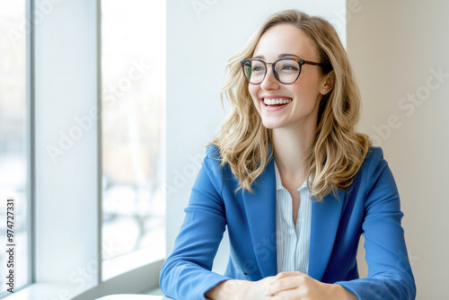 An European businesswoman in her 30s, wearing glasses, is happily smiling while sitting in her office in front of a window.