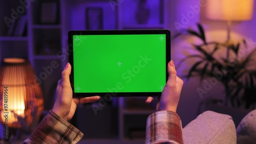 Closeup shot of girl lying on sofa using tablet in horizontal position with green screen mockup in evening room using different gestures while watching video.