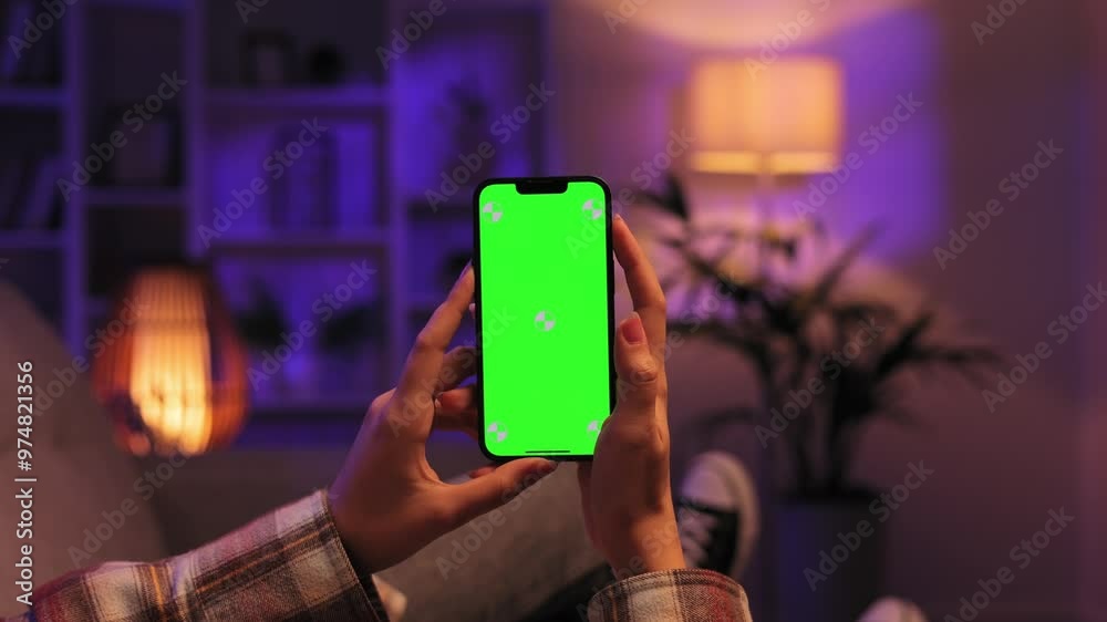 Caucasian girl scrolls sideways and taps the green screen of a smartphone with her thumb, lying on the sofa in the evening in a cozy living room. Close up of a female hand holding a mobile phone.