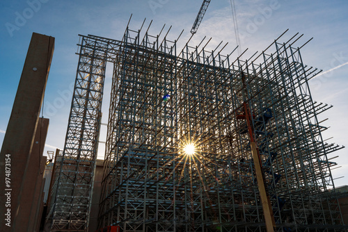 ASRS Clad rack structural framework of warehouse building skeleton during construction works with star-shaped sun behind and workers in cherry picker