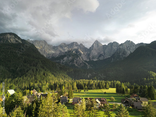 mountain village nestled at the base of towering, rugged mountains covered in dense forests. Sunlight breaks through the clouds, casting beautiful shadows over the vibrant green fields and small, scat photo