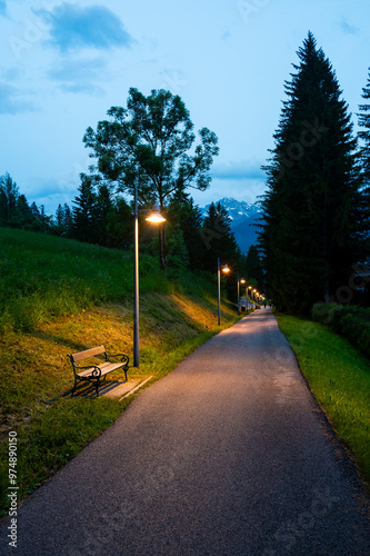 Obraz na płótnie A nighttime view of a sidewalk and cycling path illuminated by street lamps. A bench sits quietly at the edge, while the distant Dolomites are faintly visible under the night sky w salonie