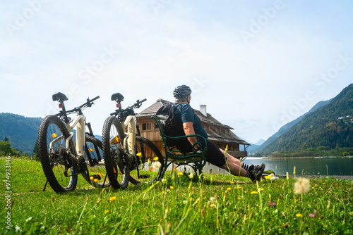 Ältere, dickerer Mann macht Pause auf einer Bank mit seinen Fahrrädern dahinter ein Holzhaus am Ossiacher See in Villach in Kärnten Österreich photo