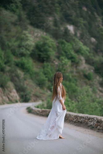 A woman in a white dress is walking down a road. The road is surrounded by trees and the woman is looking ahead. The scene has a peaceful and serene mood, as the woman is walking alone.