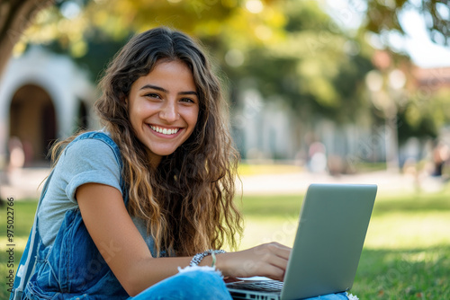 Obraz na płótnie A happy young Brazilian female college student e-learning on her laptop at campus, sitting on a lawn and looking at the camera with a big smile. w salonie