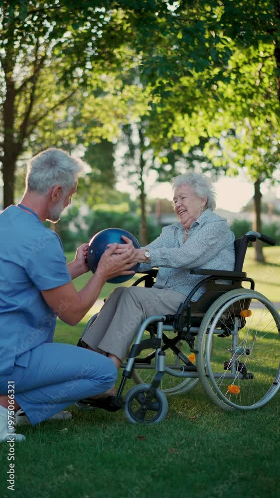 Male caregiver exercising with elderly patient on wheelchair, using small exercise ball. Geriatric physiotherapist during outdoor workout therapy in public park, nursing home garden. Male caregiver exercising with elderly patient on wheelchair, using small exercise ball. Geriatric physiotherapist during outdoor workout therapy in public park, nursing home garden.