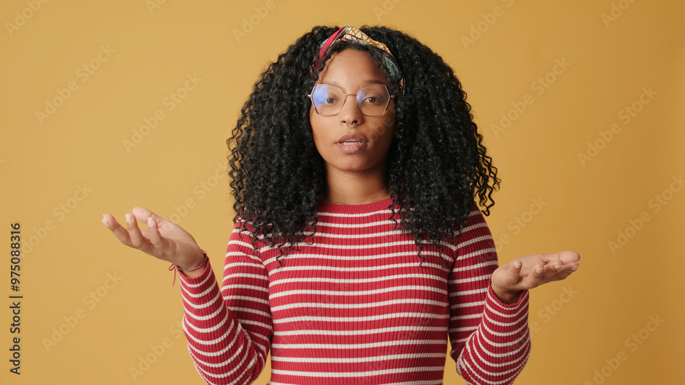 Confused young woman with curly hair wearing glasses wearing striped sweater, looking at the camera and unable to make choice, on yellow background in the studio Confused young woman with curly hair wearing glasses wearing striped sweater, looking at the camera and unable to make choice, on yellow background in the studio