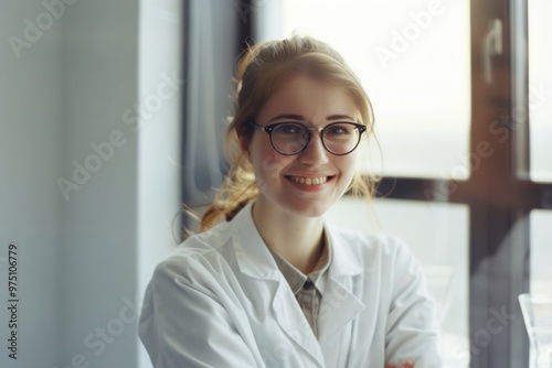 A cheerful young woman in a white lab coat, with glasses and a bright smile, captured in a sunlit room by a window.