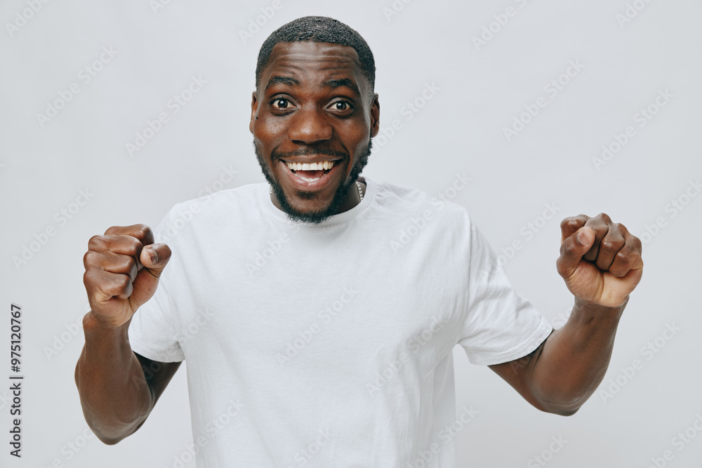 Happy young man celebrating success in a plain white t shirt against a light grey background, expressing joy and excitement with a broad smile and raised fists Happy young man celebrating success in a plain white t shirt against a light grey background, expressing joy and excitement with a broad smile and raised fists