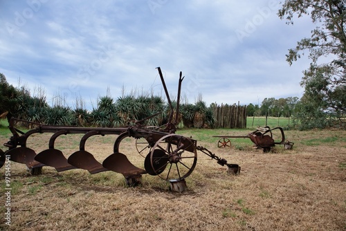 vintage plow rests against a wooden fence in a grassy field, with a backdrop of trees and a cloudy sky