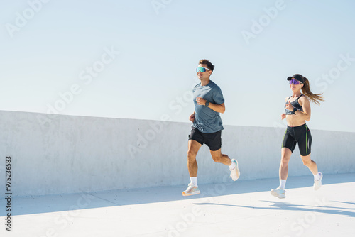 Fotografie A man and woman are jogging in a minimalist outdoor space under bright sunlight, casting long shadows on the ground