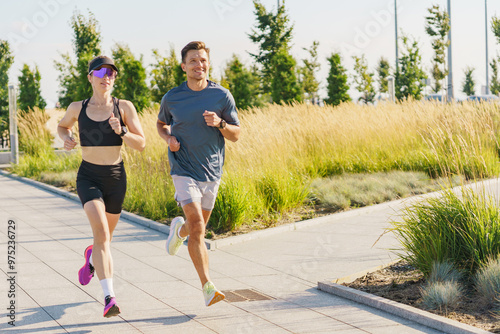 A man and woman jog along a path surrounded by tall grass and trees on a sunny day, enjoying the outdoors.