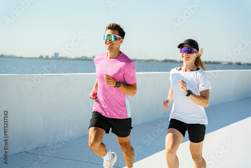 A man and woman jog together along a waterfront, smiling and enjoying the bright, sunny day, both wearing vibrant athletic wear and sunglasses. photo