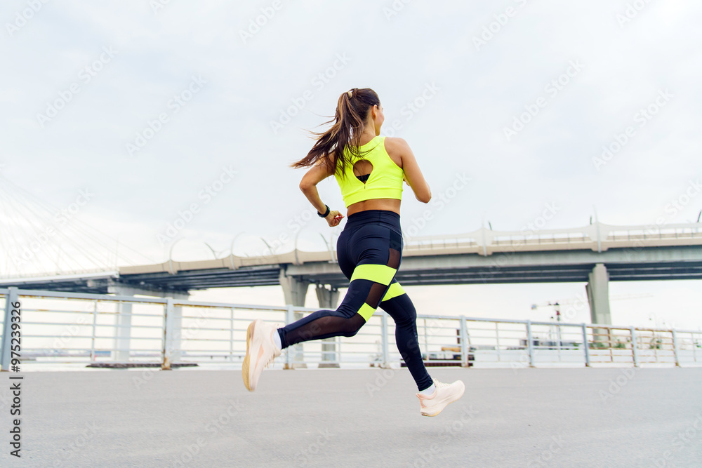 A woman in vibrant athletic wear jogs along a waterfront path, with a bridge in the background. A woman in vibrant athletic wear jogs along a waterfront path, with a bridge in the background.