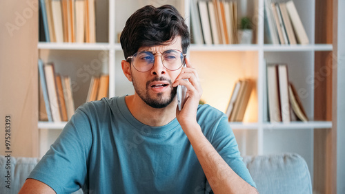 Confusing call. Mobile scam. Skeptic dissatisfied shocked puzzled man speaking on phone at modern home interior with free space.