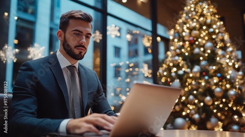 A businessman smiling while working on his laptop in a cafe, with a decorated Christmas tree and glowing lights in the background, adding a festive atmosphere.