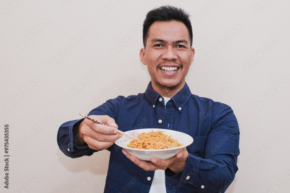 A smiling Southeast Asian man wearing casual dress holding a white plate of what instant noodles with chopsticks, the man expression is cheerful, suggesting he is proud of enjoying the meal. A smiling Southeast Asian man wearing casual dress holding a white plate of what instant noodles with chopsticks, the man expression is cheerful, suggesting he is proud of enjoying the meal.