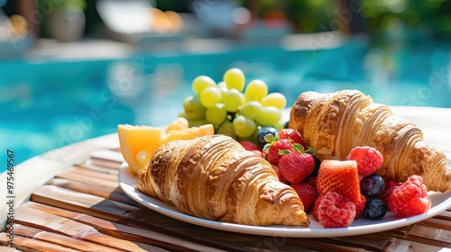 A plate of croissants and fruit is set on a table by a pool photo