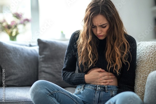 A woman is sitting on the sofa with a stomachache. Close-up shot, covering her stomach with her hand in the living room at home. This woman is feeling unwell.